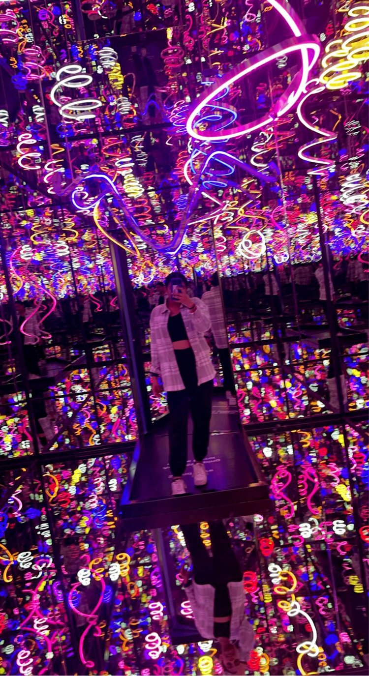 A young woman stands in a dark room lined with mirrors reflecting colorful neon curls and squiggles. She holds a phone in front of her face to take the photo.