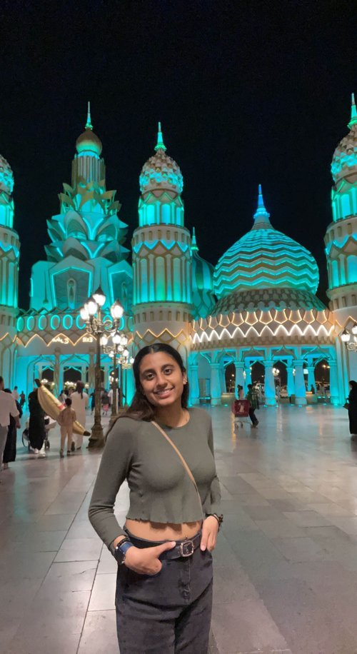 A young woman with dark features smiles at the camera, posing with her hands in her pockets. In the background is a colorfully lit architectural façade of differently styled domes and towers. It is night.