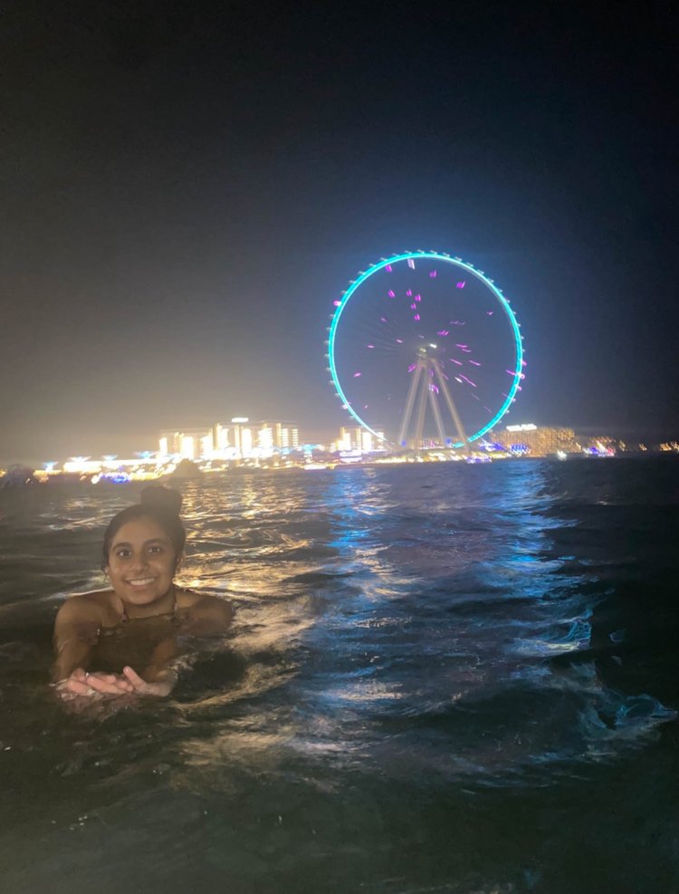 A young woman smiles at the camera. She is swimming in water up to her shoulders. It is night, and in the background is a giant lit-up Ferris wheel and some lit buildings.
