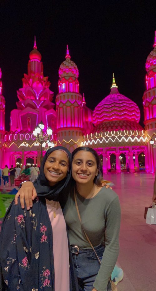 Two young women with dark features smile at the camera, posing with their arms around each other. In the background is a colorfully lit architectural façade of differently styled domes and towers. It is night.