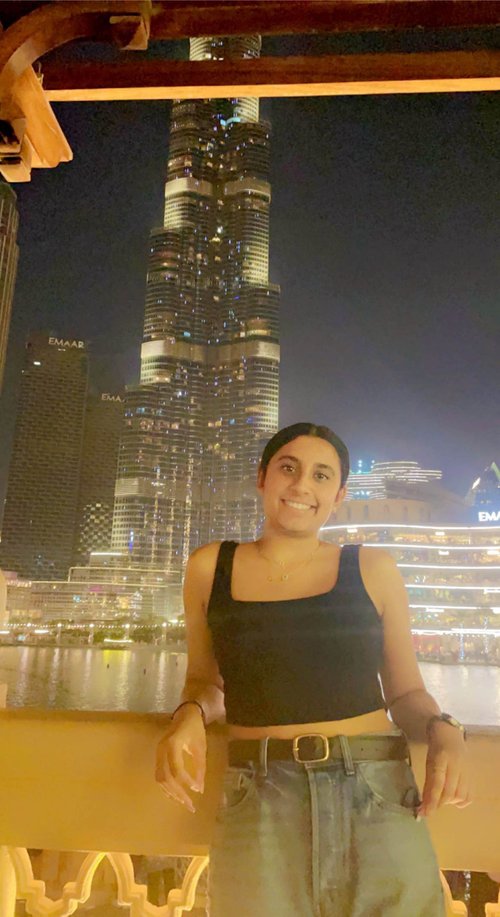 A young woman in a black top with dark features leans on a railing  around a body of water and smiles at the camera. In the background is the Burj Khalifa and other skyscrapers. It is night.
