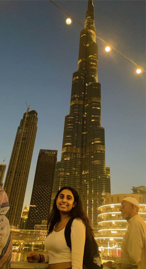 A young woman in a white top with dark features leans on a railing and smiles at the camera. In the background is the Burj Khalifa and other skyscrapers. It is night.