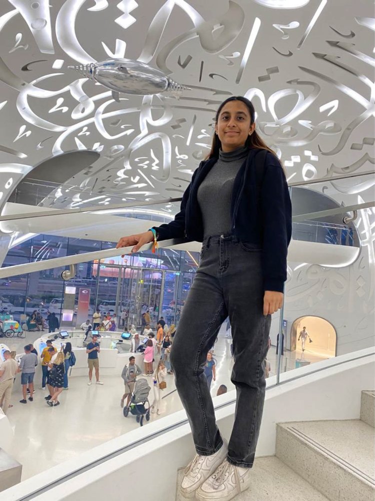 A young woman with dark features leans on a stair railing and smiles at the camera. She is standing in a museum under a silvery arched ceiling covered with giant Arabic calligraphy. There are lots of other visitors milling in the lobby.
